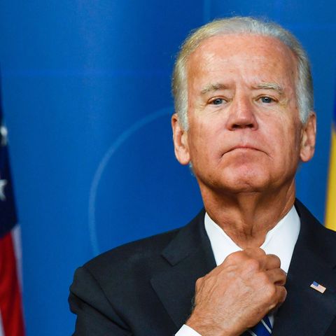 Joe Biden steht vor blaume Hintergrund und neben einer US-Flagge und richtet sich mit der rechten Hand die Krawatte