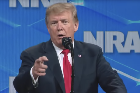 US-Präsident Donald Trump steht vor einer blauen Wand mit NRA-Logo und zielt mit dem rechten Zeigefinger ins Publikum