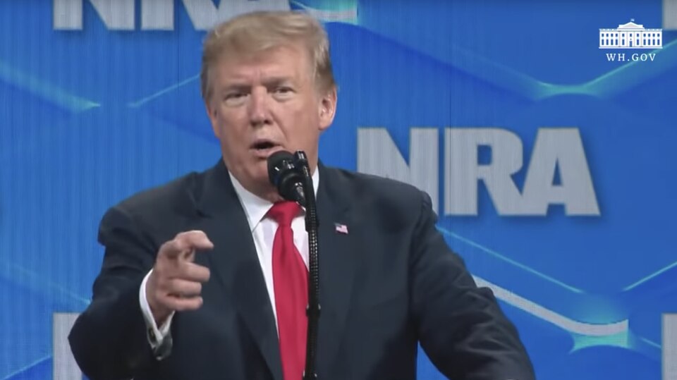 US-Präsident Donald Trump steht vor einer blauen Wand mit NRA-Logo und zielt mit dem rechten Zeigefinger ins Publikum