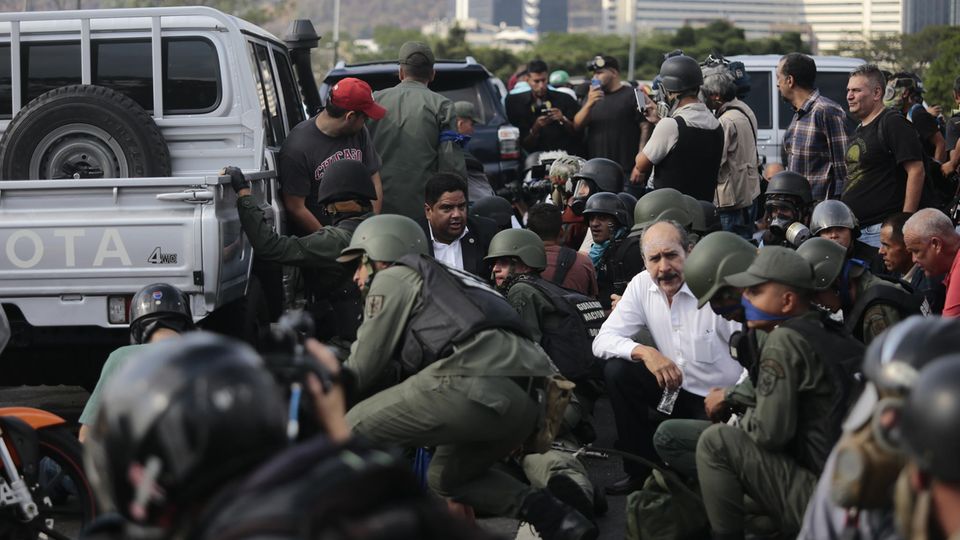 Soldaten und Zivilisten decken sich bei einem Einsatz um die Autos, in denen der selbst ernannter Interimspräsident Guaidó nahe des Luftwaffenstützpunktes La Carlota gefahren wird. "Als Interimspräsident von Venezuela, als rechtmäßiger Oberkommandierender der Streitkräfte, rufe ich alle Soldaten dazu auf, sich uns anzuschließen", sagte der Oppositionsführer auf einer Schnellstraße