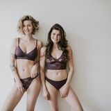 Zwei Frauen stehen in lila Unterwäsche vor einer weißen Wand.