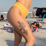 Tattoo auf dem Oberschenkel einer jungen Frau am Strand zeigt eine AK-47