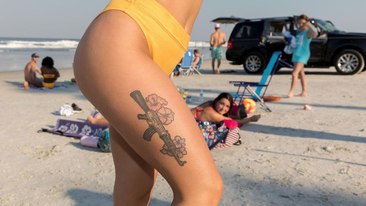Tattoo auf dem Oberschenkel einer jungen Frau am Strand zeigt eine AK-47