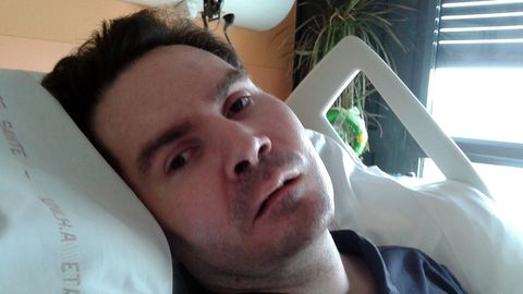 Eine Mann Ende 30 liegt in einem Krankenhausbett. Sein Kopf mit braunem Haar ist in die Kamera gedreht