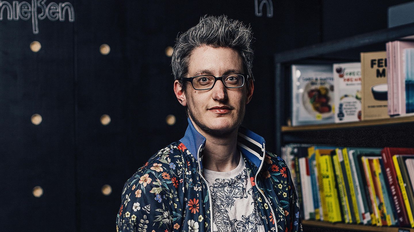 Linus Giese hat grau-schwarze Haare, trägt eine Brille und hat Bartwuchs. Er trägt ein Shirt und eine Jacke mit Blumenmustern