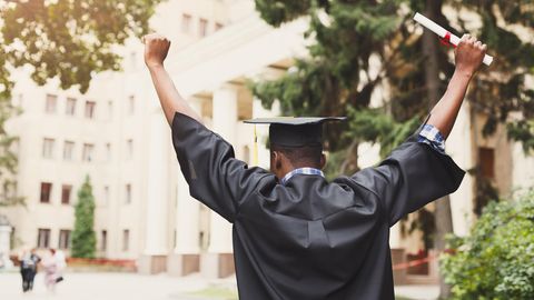 Ein junger, schwarzer Schüler von hinten. Er trägt eine Abschlussrobe, einen Uni-Abschluss-Hut und hält ein Diplom in der Hand