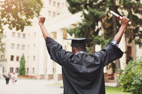 Ein junger, schwarzer Schüler von hinten. Er trägt eine Abschlussrobe, einen Uni-Abschluss-Hut und hält ein Diplom in der Hand