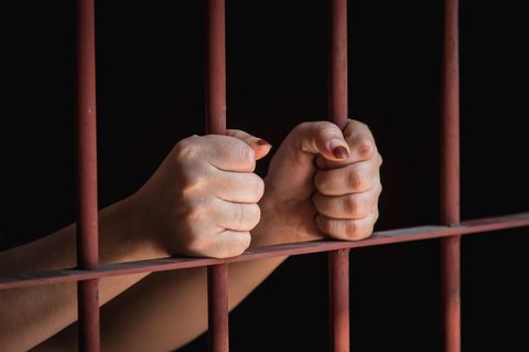 Frauenhände mit halb abgesplittertem Nagellack umklammern rote Gefängnisstäbe