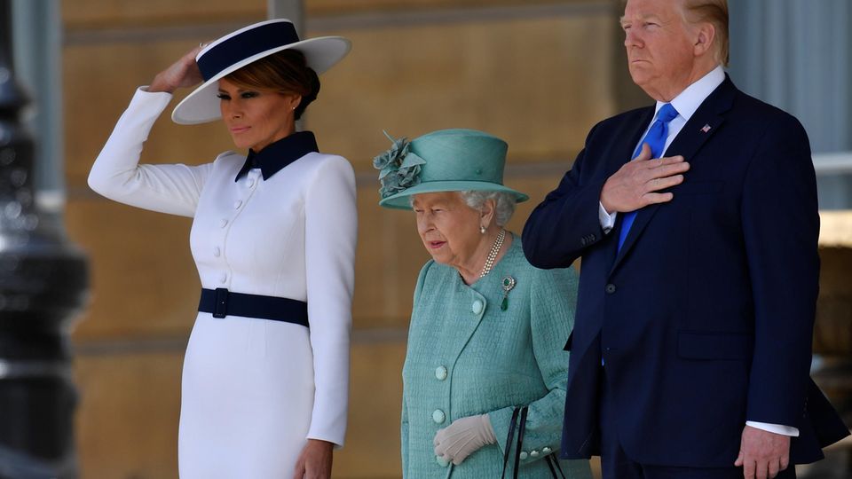Die Hand gehört eigentlich nicht an den Hut: Melania Trump (l.) muss bei einem Windstoß nach oben greifen. Der Hut der Queen sitzt hingegen perfekt. Donald Trump bleibt unbeirrt.
