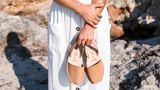 Frau in weißem Kleid hält ein Paar Espadrilles in der Hand.