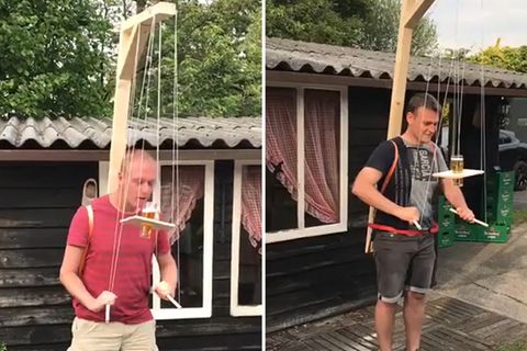 Mit Hilfe eines marionettenartigen Gestells müssen Männer ein Bierglas auf einem Holzbrett zum Mund balancieren.
