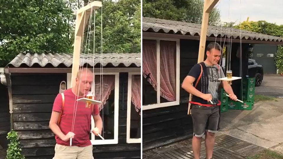 Mit Hilfe eines marionettenartigen Gestells müssen Männer ein Bierglas auf einem Holzbrett zum Mund balancieren.