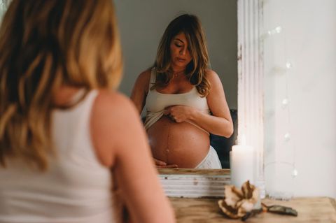 Eine junge, schwangere Frau steht vor dem Spiegel und schaut auf ihren Bauch. Darauf sieht man einen vertikalen Streifen