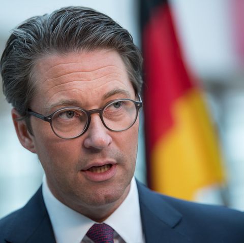 Ein Mann mit braunem Seitenscheitel und runder Brille steht mit offenem Mund im Anzug vor einer Deutschland-Flagge