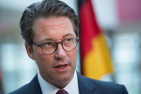 Ein Mann mit braunem Seitenscheitel und runder Brille steht mit offenem Mund im Anzug vor einer Deutschland-Flagge