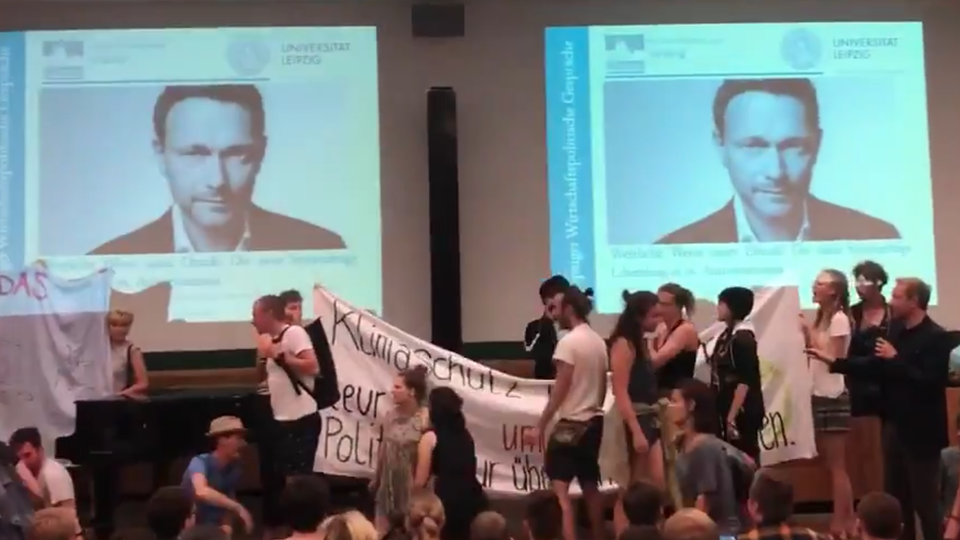 Auf einer Bühne stehen junge Menschen mit Bannern, im Hintergrund sind zwei Porträts von Christian Lindner projeziert