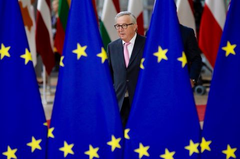 Ein Mann mit grauem Seitenscheitel und Brille läuft im Anzug zwischen Europa-Flaggen hindurch