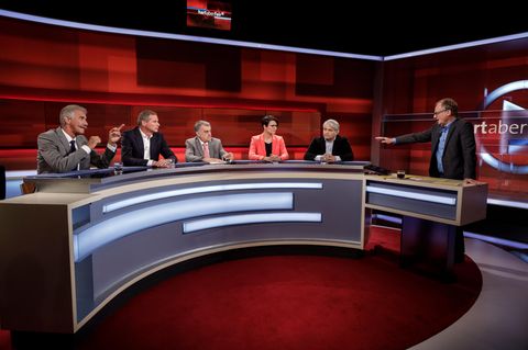 In einem Fernsehstudio mit rötlichem Hintergrung sitzen an einem halbrunden Tisch vier Männer und eine Frau. Rechts steht einer