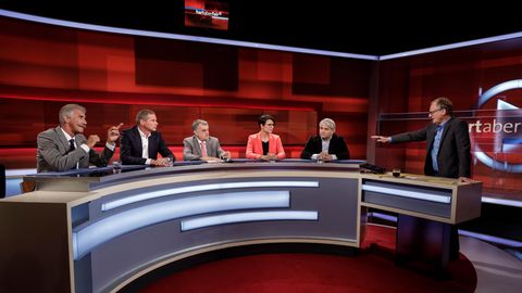 In einem Fernsehstudio mit rötlichem Hintergrung sitzen an einem halbrunden Tisch vier Männer und eine Frau. Rechts steht einer