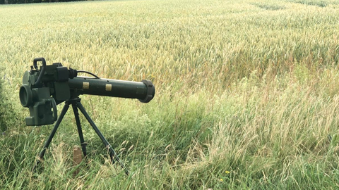 Neben dem Stamm einer Birke steht ein Raketenwerfer auf einem dreibeinigen Stativ im Gras am Feldrand