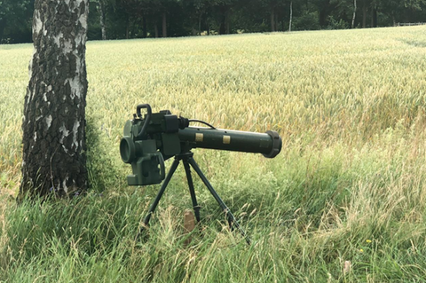 Neben dem Stamm einer Birke steht ein Raketenwerfer auf einem dreibeinigen Stativ im Gras am Feldrand
