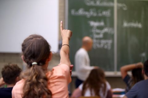 Ein Mädchen meldet sich im Klassenzimmer, im Hintergrund ist ein Lehrer zu sehen