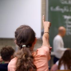 Ein Mädchen meldet sich im Klassenzimmer, im Hintergrund ist ein Lehrer zu sehen