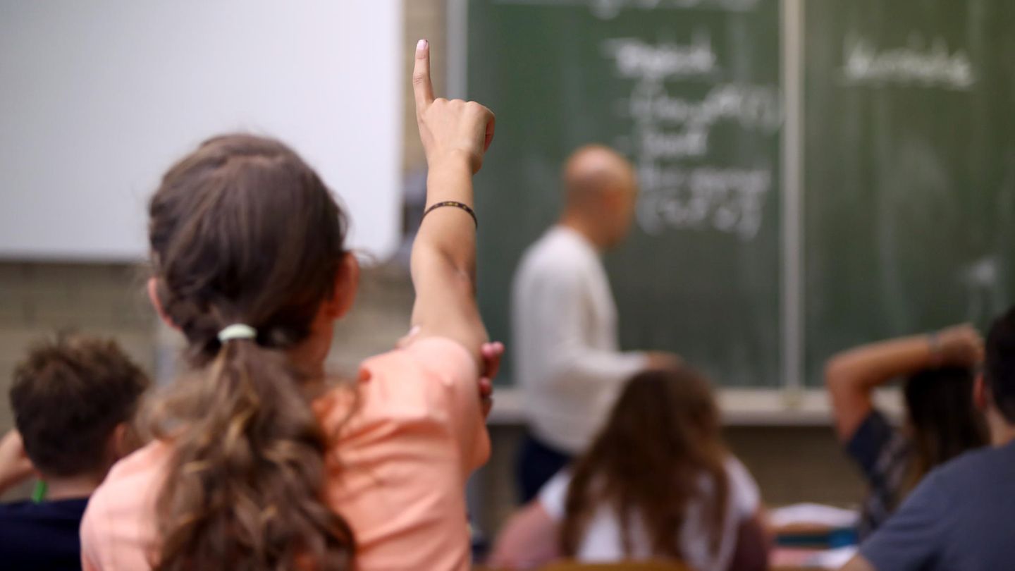 Ein Mädchen meldet sich im Klassenzimmer, im Hintergrund ist ein Lehrer zu sehen
