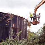 Zwei junge Männer stehen auf der Schaufel eines Baggers in luftiger Höhe
