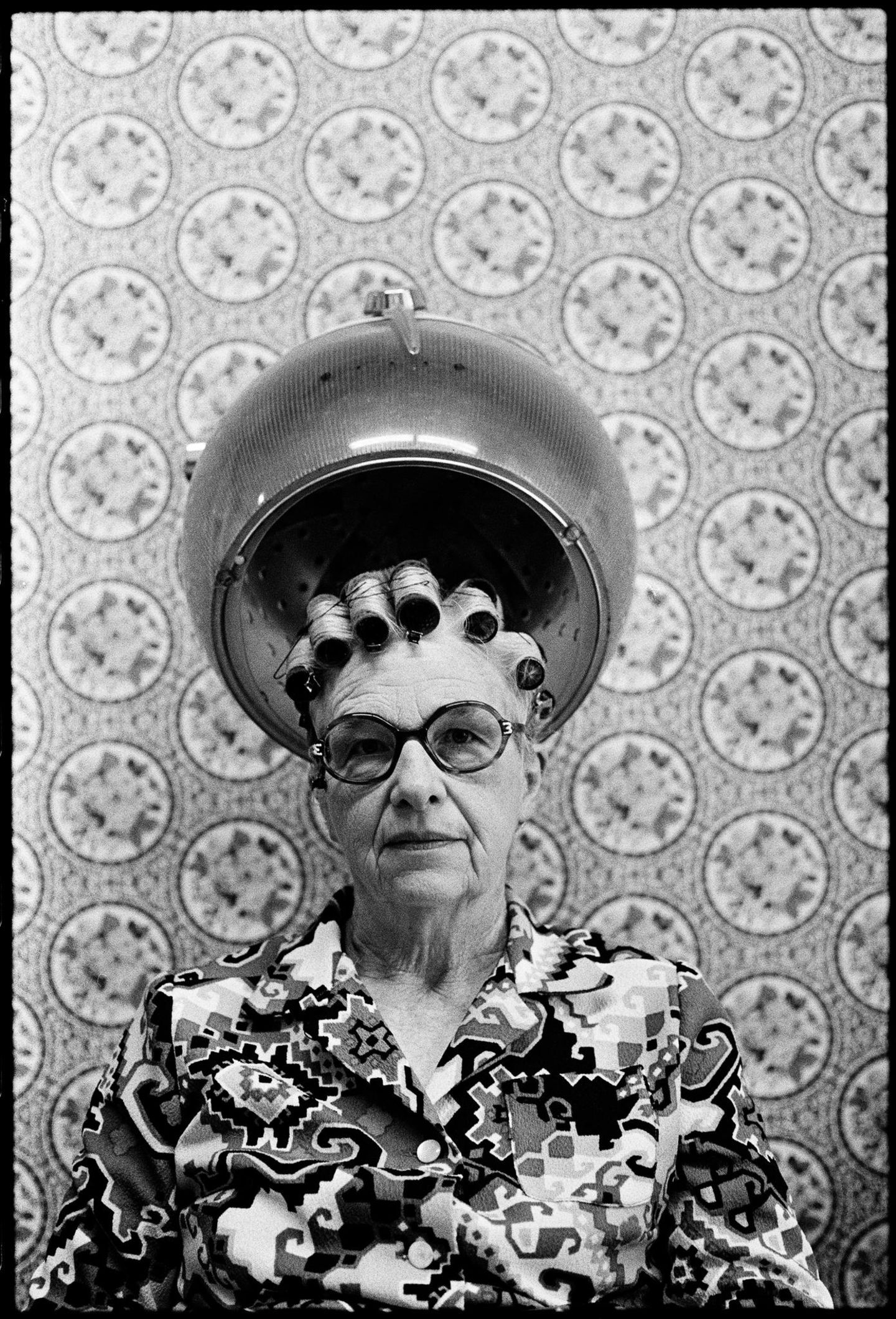 Eine alte Frau mit Brille sitzt unter einer Trockenhaube