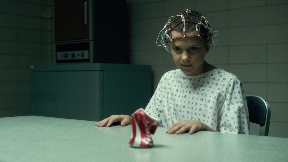 Elfie aus "Stranger Things" sitzt an einem Tisch mit Elektroden am Kopf und zerquetscht mit ihren Gedanken eine Coca-Cola-Dose