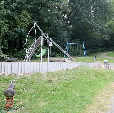 Spielgeräte stehen vor Bäumen. Der Spielplatz ist mit Betonpollern abgegrenzt. Ein Sandweg führt über eine Wiese daran vorbei
