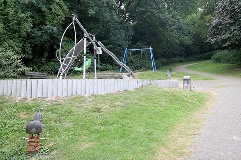 Spielgeräte stehen vor Bäumen. Der Spielplatz ist mit Betonpollern abgegrenzt. Ein Sandweg führt über eine Wiese daran vorbei