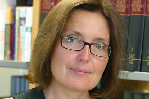Eine Frau mit schmaler Brille und schulterlangen Haaren steht mit nach links geneigtem Kopf vor einem Bücherregal