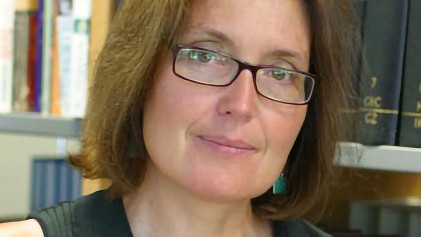 Eine Frau mit schmaler Brille und schulterlangen Haaren steht mit nach links geneigtem Kopf vor einem Bücherregal