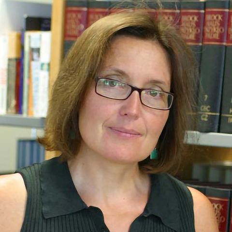 Eine Frau mit schmaler Brille und schulterlangen Haaren steht mit nach links geneigtem Kopf vor einem Bücherregal