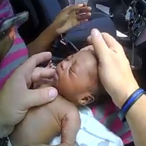 Links hält eine rechte Hand den Kopf eines Babys, eine linke fühlt dessen Hals. Rechts steht ein US-Polizist neben einer US-Flag
