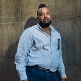 Ein schwarzer Mann mit Irokesenhaarschnitt lehnt an einer Wand