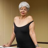 Mit dichtem weißen Haar und einem weit ausgeschnittenen Kleid stützt sich eine ältere Dame auf ein Geländer