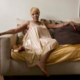 Eine schwarze Frau mit blonder Perücke und langen roten Fingernägeln sitzt auf einem Sofa