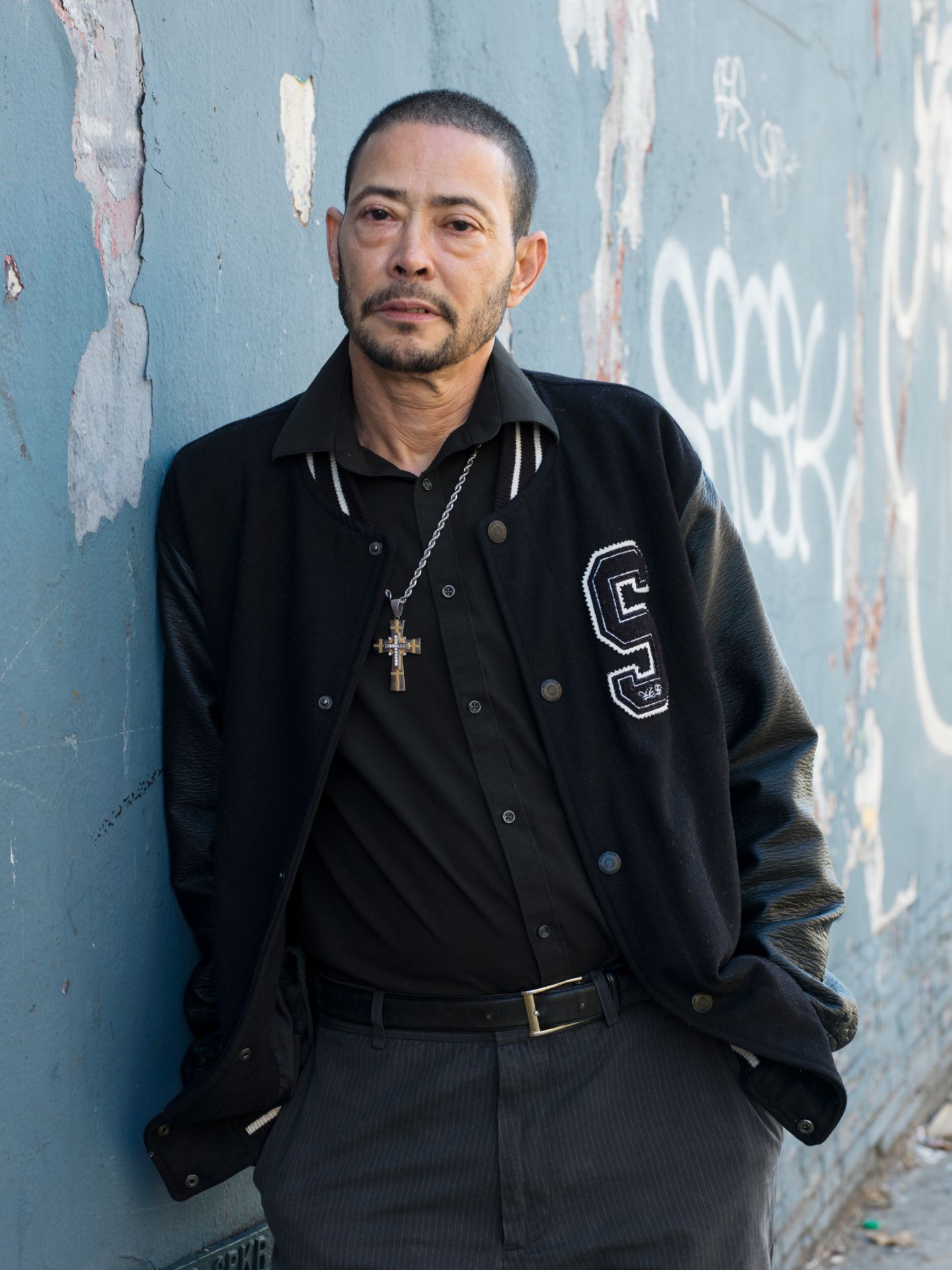 Ein älterer hispanischer Mann in Collegejacke und kurz geschorenem Haar lehnt an einer Wand