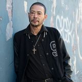 Ein älterer hispanischer Mann in Collegejacke und kurz geschorenem Haar lehnt an einer Wand