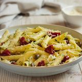 Nudelsalat mit getrockneten Tomaten  Zutaten:  Für die halb getrockneten Tomaten: 1 kg Cocktailtomaten, 1 EL Zucker, Meersalz, schwarzer Pfeffer aus der Mühle, 1 EL Thymianblättchen, 4 EL natives Olivenöl extra  Für den Salat: 500 g kurze Nudeln (z. B. Fusilli), Meersalz, 50 g Pinienkern, 2 Kugeln Büffelmozzarella (à 125 g Abtropfgewicht), 60 g Parmesan, 1 großes Bund Basilikum, 150 g Rucola  Für die Sauce: 6 EL Aceto balsamico, 4 EL Zitronensaft, 1 EL Akazienhonig, 100 ml Gemüsebrühe, Meersalz, schwarzer Pfeffer aus der Mühle, 8 EL natives Olivenöl extra  Zubereitung: Am Vorabend den Backofen auf 200 °C (Umluft 180 °C) vorheizen. Die Tomaten waschen, trocken reiben und halbieren. Ein Backblech mit Backpapier belegen und die Tomaten mit der Schnittfläche nach oben darauf verteilen. Zucker, 2 Teelöffel Meersalz, reichlich Pfeffer und die Thymianblättchen mischen und über die Tomaten streuen. Mit Olivenöl beträufeln. Das Backblech in den heißen Ofen schieben und die Tomaten 20 Minuten garen. Dann den Ofen ausschalten und die Tomaten über Nacht in der Resthitze des Backofens trocknen lassen.  Am nächsten Tag die Nudeln in reichlich Salzwasser bissfest kochen, dann durch ein Sieb abgießen, unter fließendem kaltem Wasser abschrecken und abtropfen lassen.  Pinienkerne in einer Pfanne ohne Fett goldbraun rösten, leicht salzen und beiseite stellen. Den Mozzarella abtropfen lassen und in kleine Stücke zupfen. Den Parmesan grob reiben oder in kleine Stücke brechen. Basilikum waschen und trocken schütteln. Die Blätter von den Stängeln zupfen und etwas kleiner zupfen. Den Rucola verlesen, waschen, trocken schleudern und in einen Gefrierbeutel füllen. Den Beutel verschließen und bis zum Servieren im Kühlschrank aufbewahren.  Für die Sauce Aceto balsamico, Zitronensaft, Honig, Gemüsebrühe, etwas Meersalz, reichlich Pfeffer und das Olivenöl kräftig verrühren. Nudeln, Tomaten, Mozzarella, Parmesan und Basilikum mit der Sauce in einer großen Schüssel mischen und mindestens 30 Minuten ziehen lassen.  Zum Servieren den Salat durchmischen und bei Bedarf nachwürzen. Dann den Rucola unterheben und den Salat anrichten. Mit den Pinienkernen bestreuen.