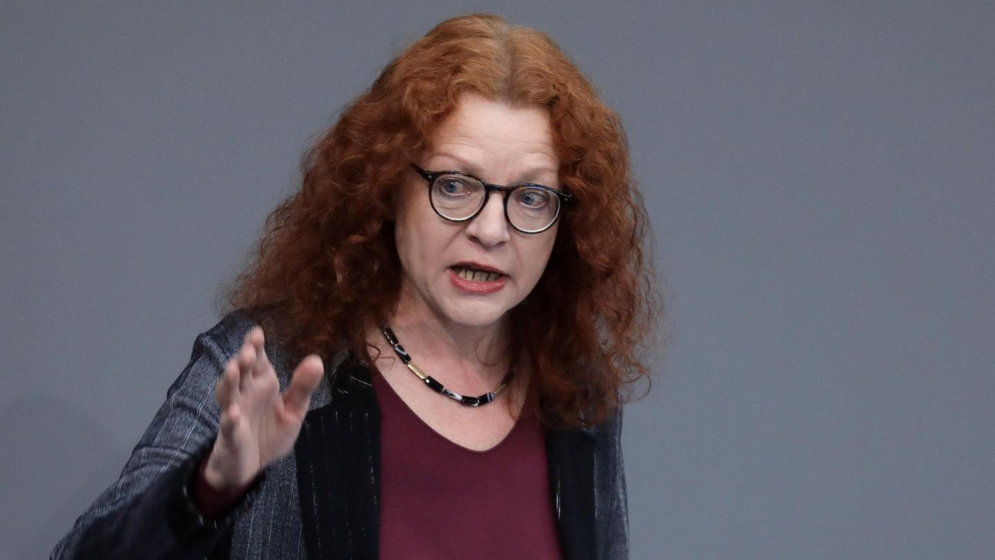 Eine Frau mit langen roten Locken gestikuliert mit der rechten Hand vor einem grauen Hintergrund