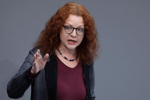 Eine Frau mit langen roten Locken gestikuliert mit der rechten Hand vor einem grauen Hintergrund