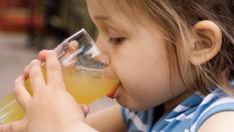 Ein dunkelblondes kleines Mädchen führt mit beiden Händen ein Glas Apfelsaft zum Mund