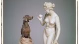 Ein Weimaraner sitzt auf einem Sockel und blickt in die Augen einer Statue, die einen Ball in Baseballgröße hält