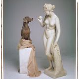 Ein Weimaraner sitzt auf einem Sockel und blickt in die Augen einer Statue, die einen Ball in Baseballgröße hält