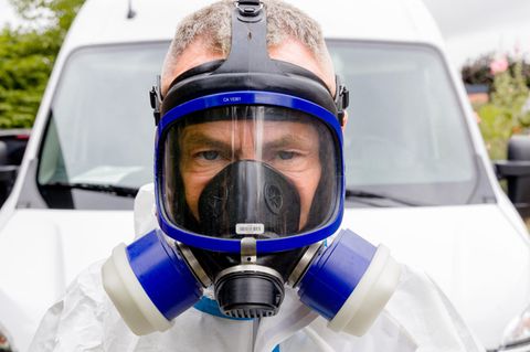 Ein Mann mit blauer Gasmaske und weißem Schutzanzug steht vor einem weißen Auto und schaut in die Kamera