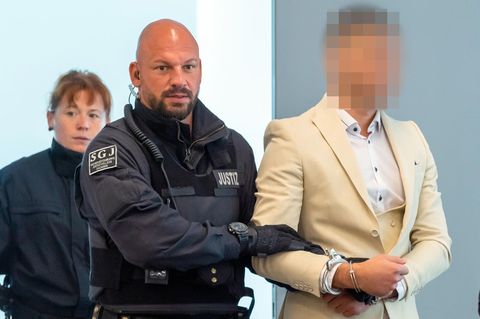 Ein Mann im weißen Anzug und Hemd wird von einem kahlköpfigen Polizisten mit Bart abgeführt